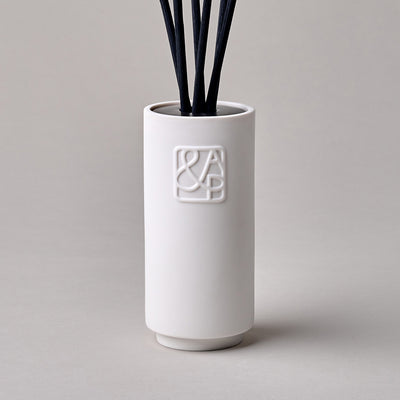 A white ceramic diffuser with black rattan reeds, featuring a logo on the front, designed to complement any space.
