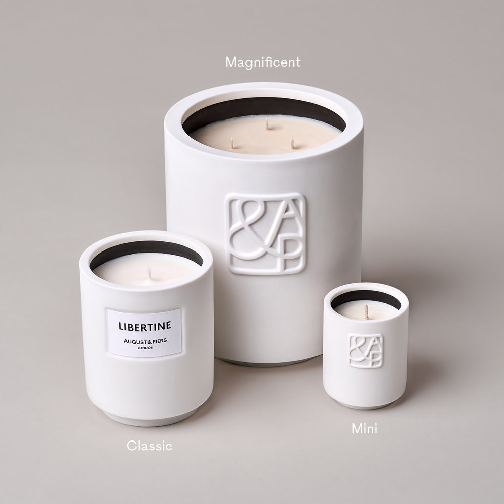 Three white candles of varying sizes labeled as 'Magnificent,' 'Classic,' and 'Mini.' The 'Classic' candle features the label 'LIBERTINE, AUGUST & PIERS, LONDON,' while the 'Magnificent' and 'Mini' candles showcase an embossed 'A&P' logo, set against a neutral background.