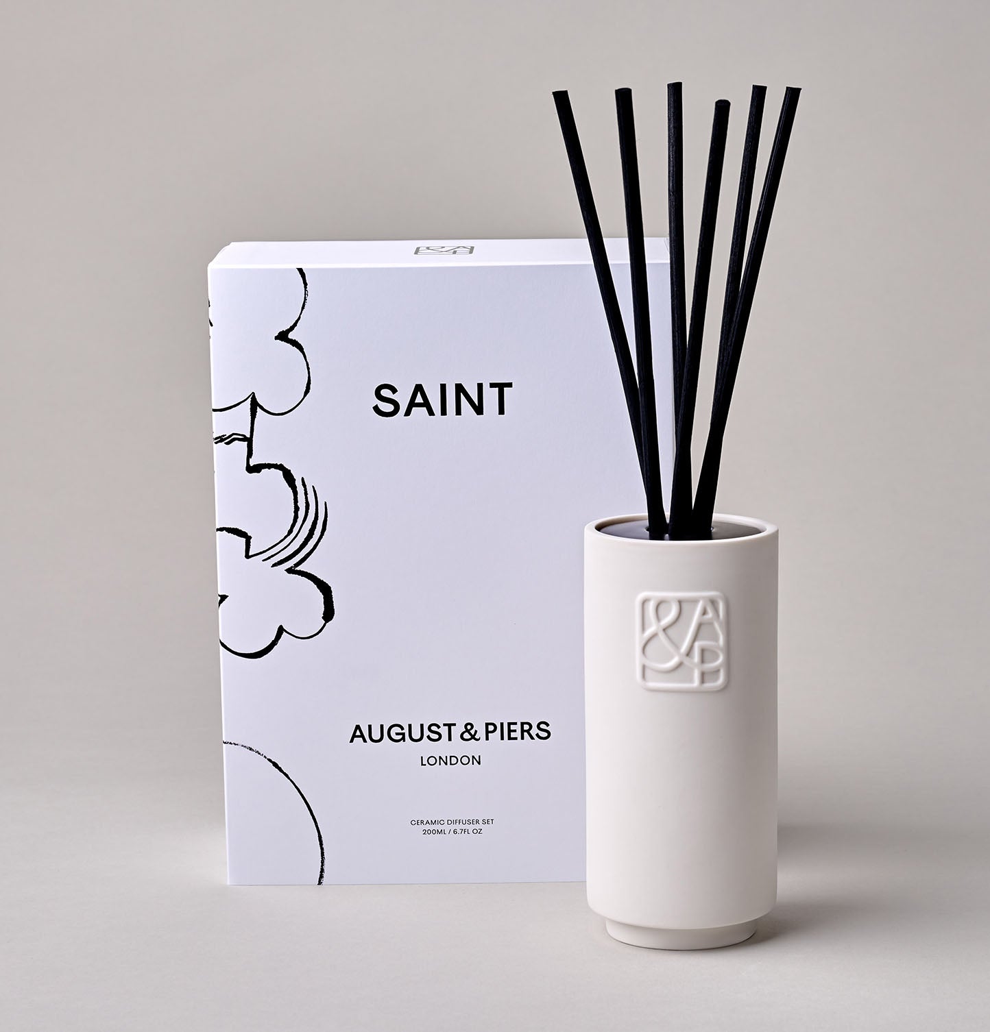 AUGUST&PIERS white ceramic diffuser with embossed 'A&P' logos on the front, displayed alongside their packaging box featuring an illustration by Petra Börner, set against a neutral background.