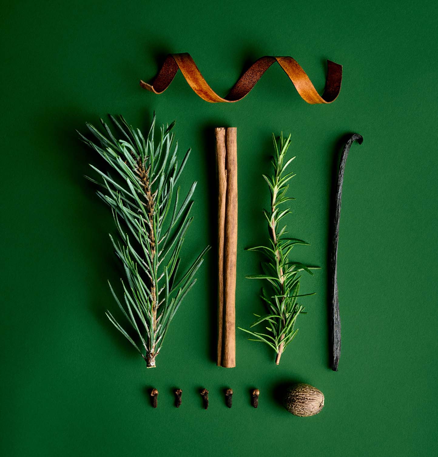 Flat lay of rosemary sprigs, cinnamon stick, vanilla pod, clove buds, nutmeg and orange peel on a green background — ingredients evoking the festive fragrance notes of AUGUST&PIERS Claus candle.