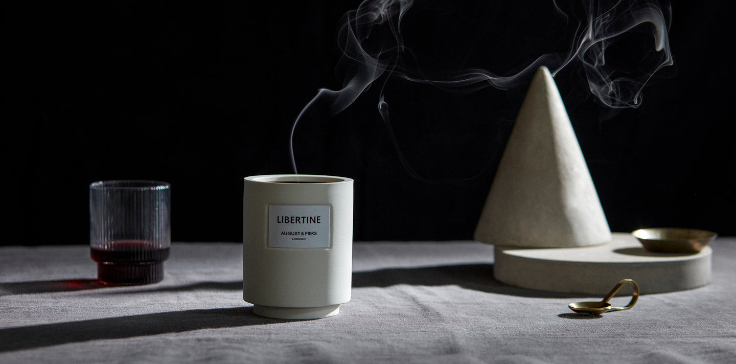 Libertine luxury fragranced candle smoking on a grey linen cloth table top with dramatic lighting.