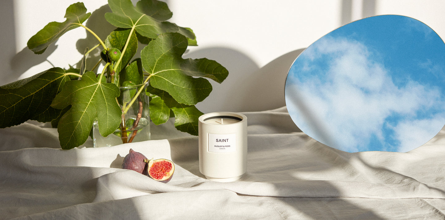 Saint luxury fragranced candle situated on linen cloth table next to wild fig leafs and an oval mirror reflecting the sky.
