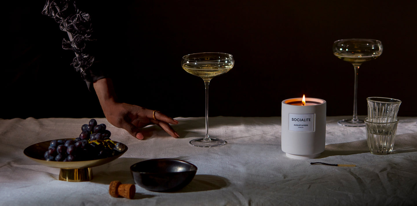 Socialite luxury fragranced candle situated on a grey cloth table surrounded by champagne flutes and black grapes in a brass dish.